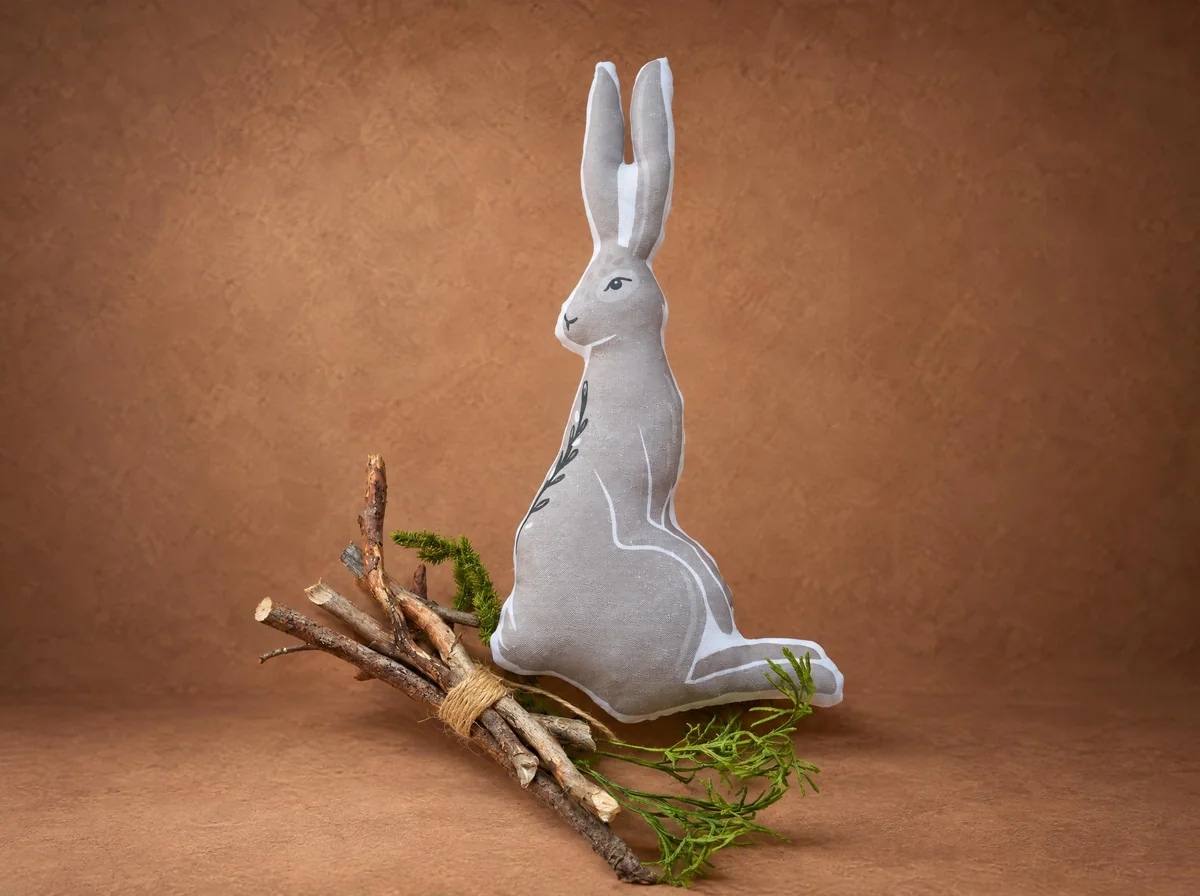 Подушка-заяц в прыжке из серой ткани, сшита по рисунку для коллекции Ginger Bird, рядом ветки и зелень